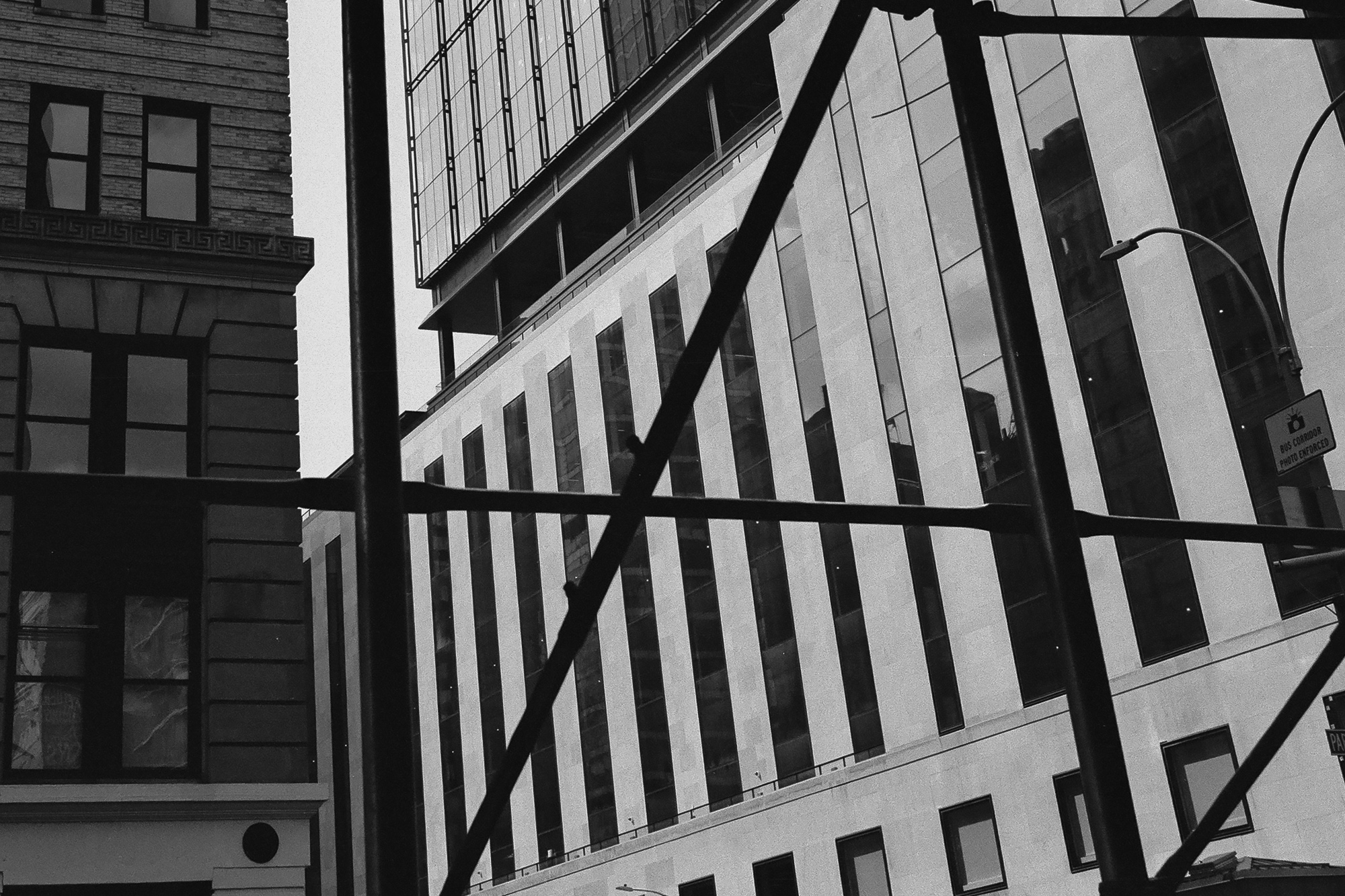 Black and white street photograph looking up through dark scaffolding pipes that cross the frame in vertical, horizontal and diagonal lines, framing a modern tower with alternating stone piers and dark glass windows, with an older brick building at left and a curved streetlight at right.