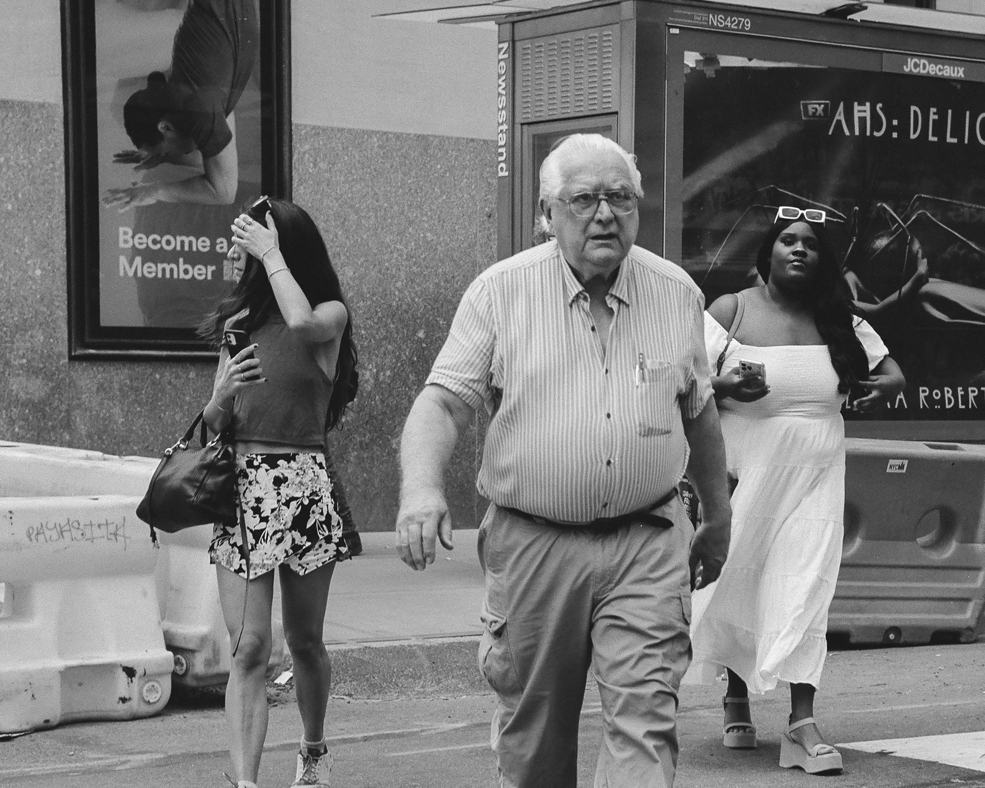 Black and white street photograph of an older man with white hair, glasses and a striped short-sleeve shirt walking across a street, flanked by a young woman in a floral skirt holding a phone and pushing her hair back and a woman in a white dress with sunglasses on her head. Behind them stand a newsstand kiosk and a large billboard, with concrete traffic barriers at the curb.