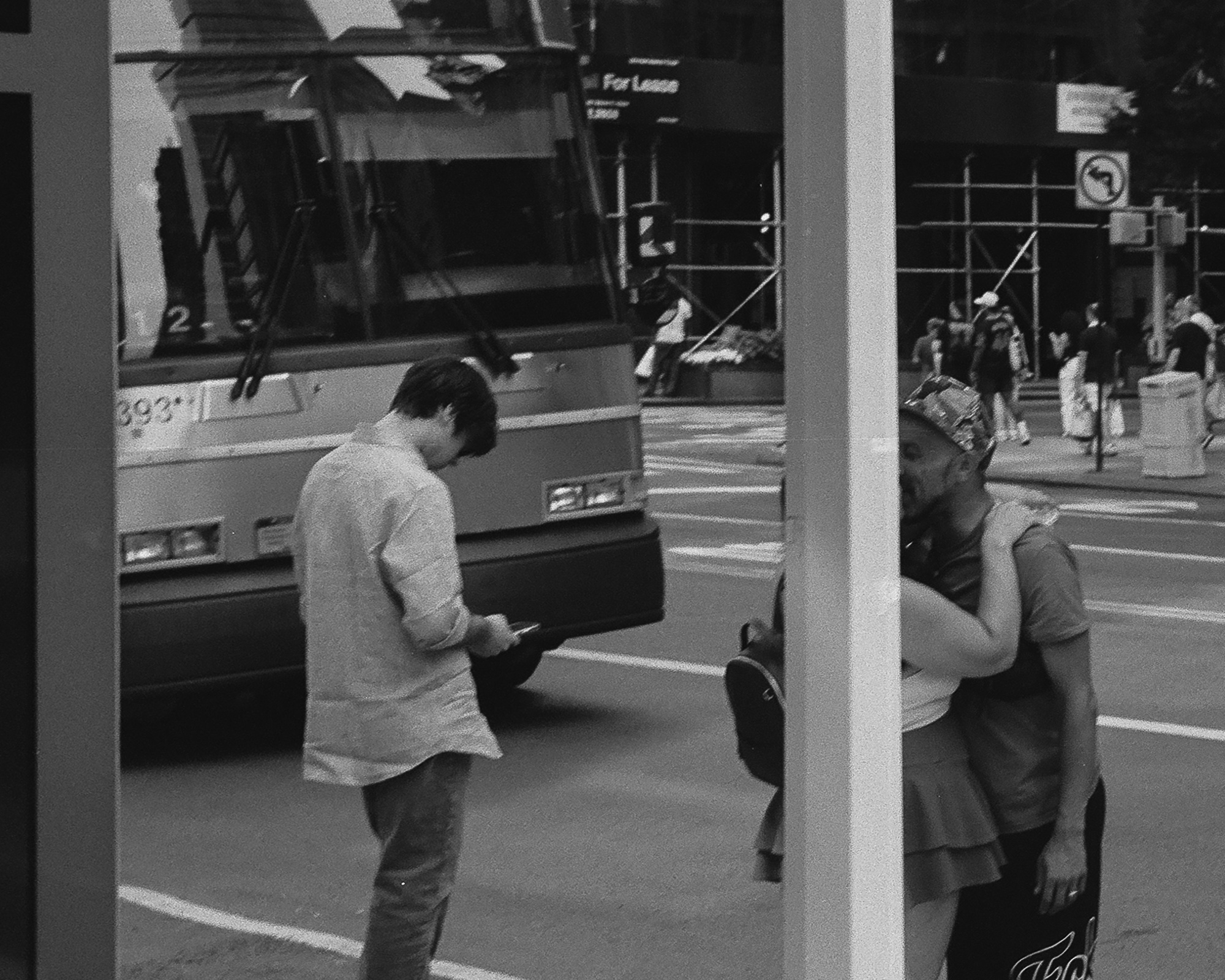 Black and white street photograph of a Manhattan corner: at left a young man in a light untucked shirt stares down at his phone as a city bus passes behind him, while at right, beyond a white vertical pole dividing the frame, a couple stand locked in an embrace, her hand on his shoulder beneath a patterned cap.