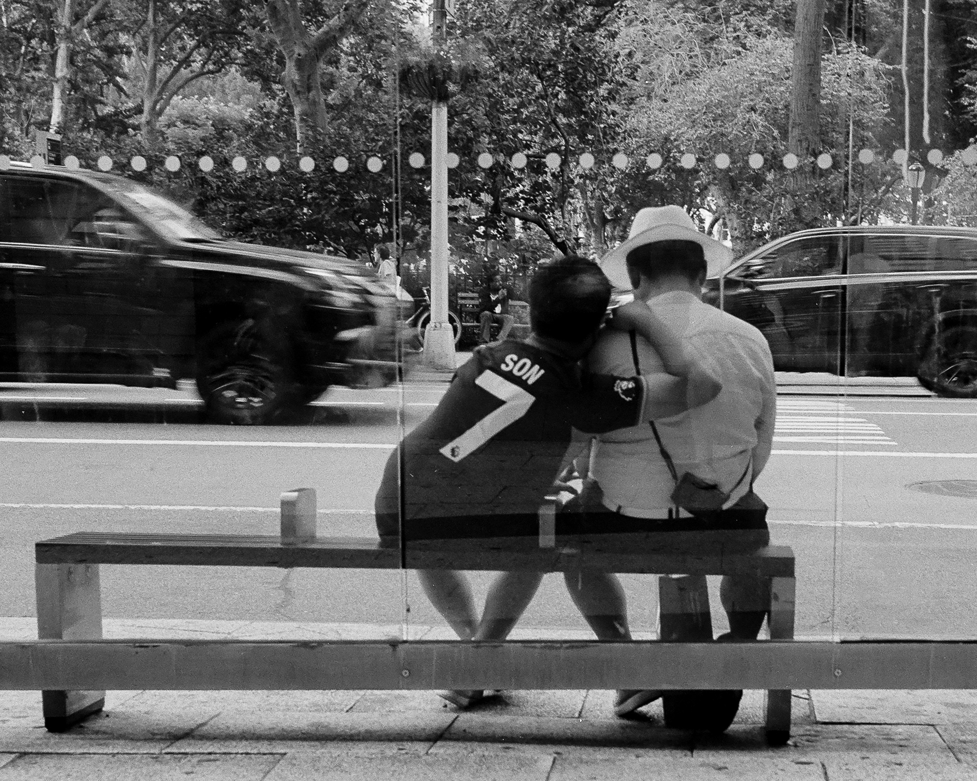 Black and white photo of two people seen from behind, seated close together on a bus shelter bench. One wears a dark soccer jersey printed SON and the number 7; the other wears a wide white sun hat and a light shirt, one arm around the first person's shoulder. Behind them, a dark SUV blurs past on the street and park trees fill the background through the shelter glass.
