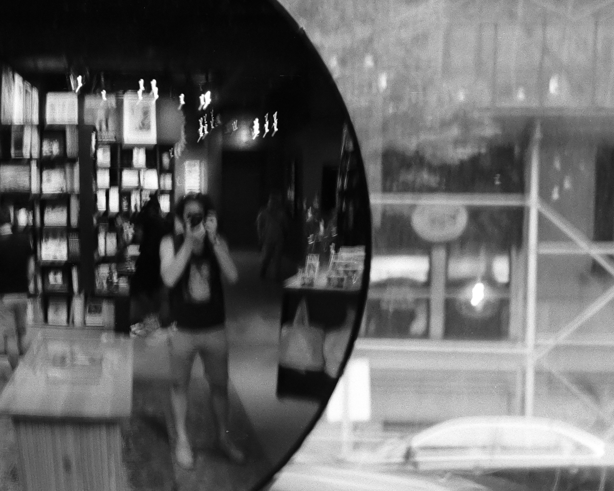Black and white photograph of a large round convex mirror mounted in a bookstore, reflecting a masked person holding a camera to their face, surrounded by bookshelves, ceiling lights and a second figure walking past. To the right of the mirror, a window shows blurred street scaffolding and sidewalk.