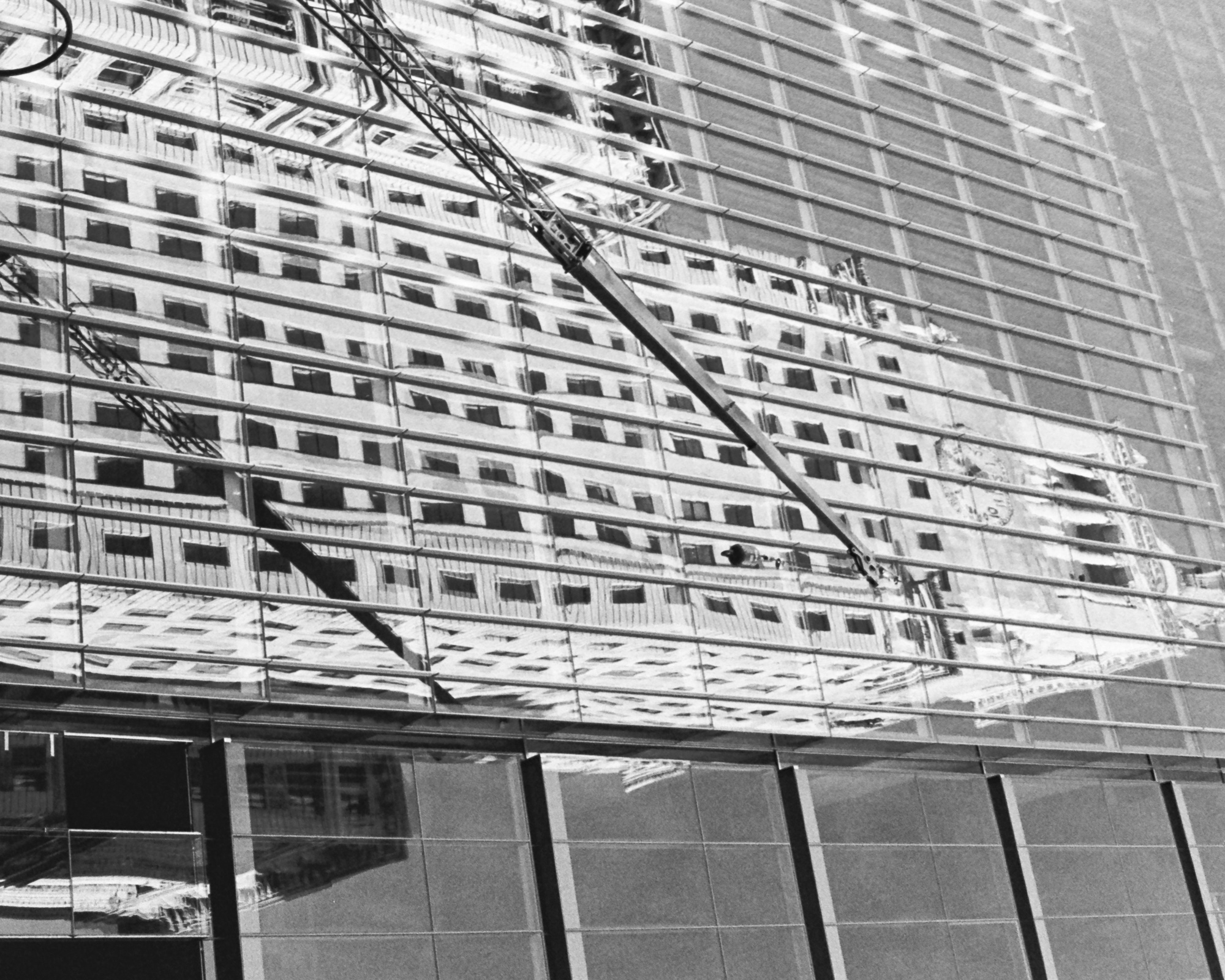 Black and white photograph of a mirrored glass office facade in New York City, its horizontal mullions slicing a reflected prewar apartment block into warped bands. A construction crane's lattice boom cuts diagonally across the reflection from upper left to lower right, ending in a hook block above the darker granite and glass of the building's lower floors.
