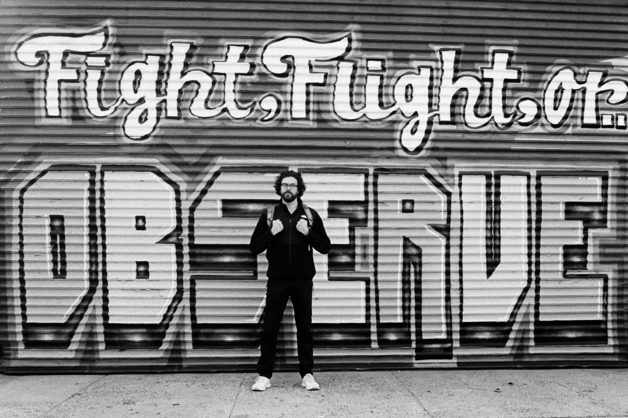 A bearded man in glasses, a dark zip jacket, dark pants and white sneakers stands facing the camera on a sidewalk, gripping his backpack straps, in front of a corrugated metal shutter painted with the words 'Fight, Flight, or...' above giant block letters spelling 'OBSERVE.' The photograph is black and white.