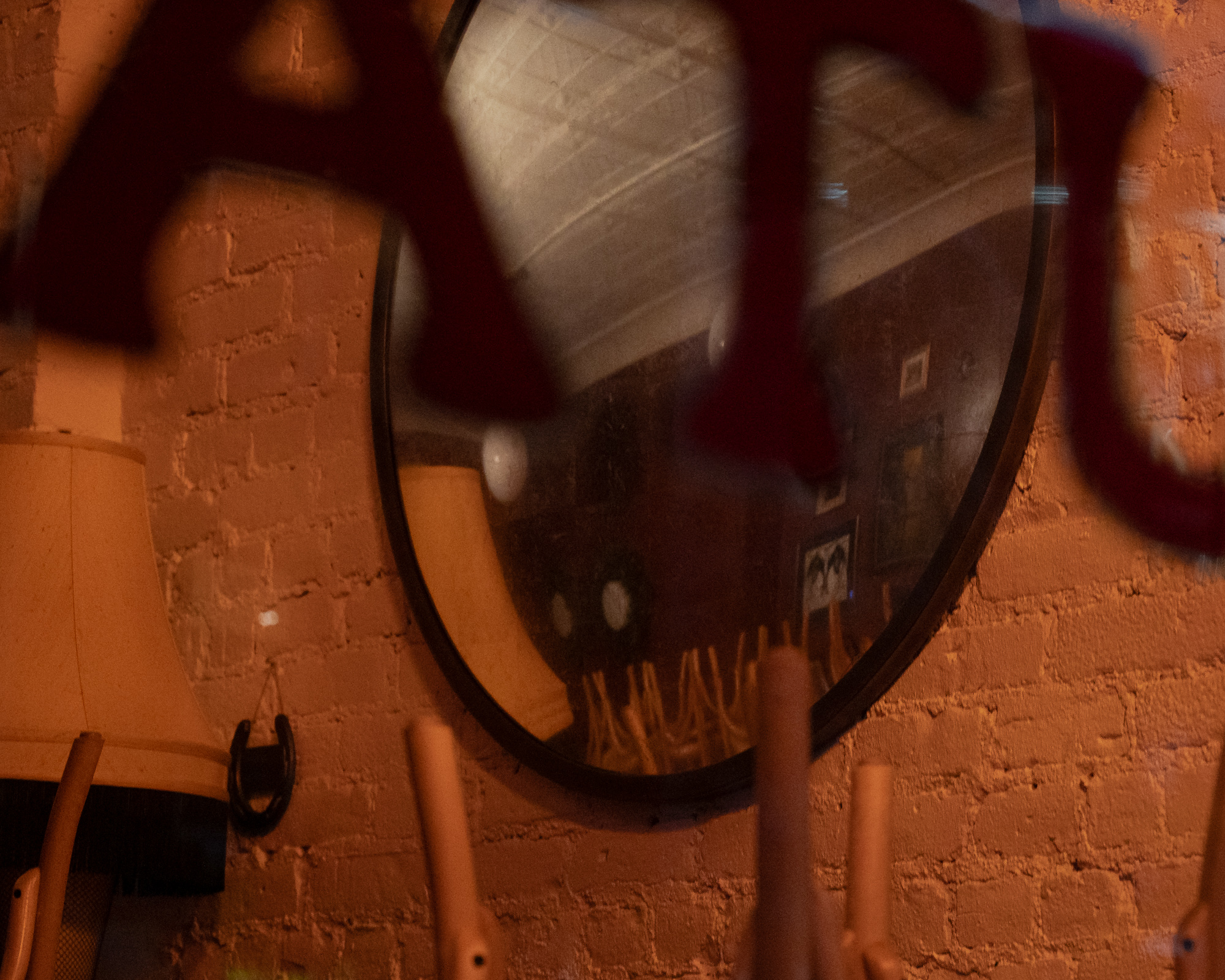An oval mirror hangs on an orange painted brick wall, reflecting a dim bar interior with a pressed tin ceiling, framed pictures and a chandelier. A lampshade and an iron horseshoe hang on the wall nearby, while blurred dark red shapes drift across the foreground.