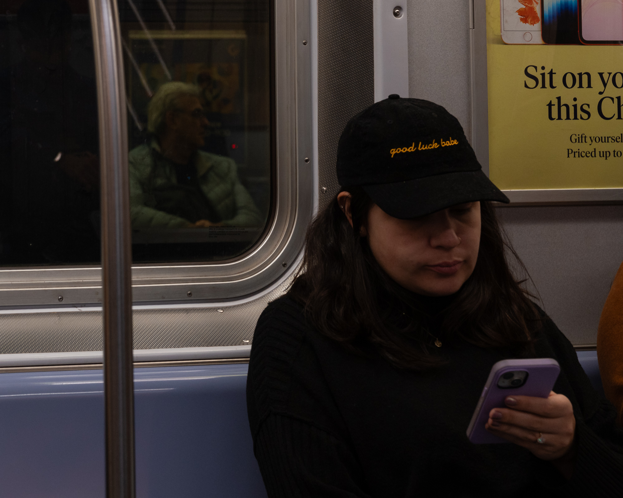 A woman in a black ribbed sweater and a black baseball cap embroidered with the words 'good luck babe' in orange script looks down at a lavender smartphone on a subway car. In the window behind her, the reflection of an older man in a pale green puffer jacket sits facing the other direction, beside a yellow advertisement.