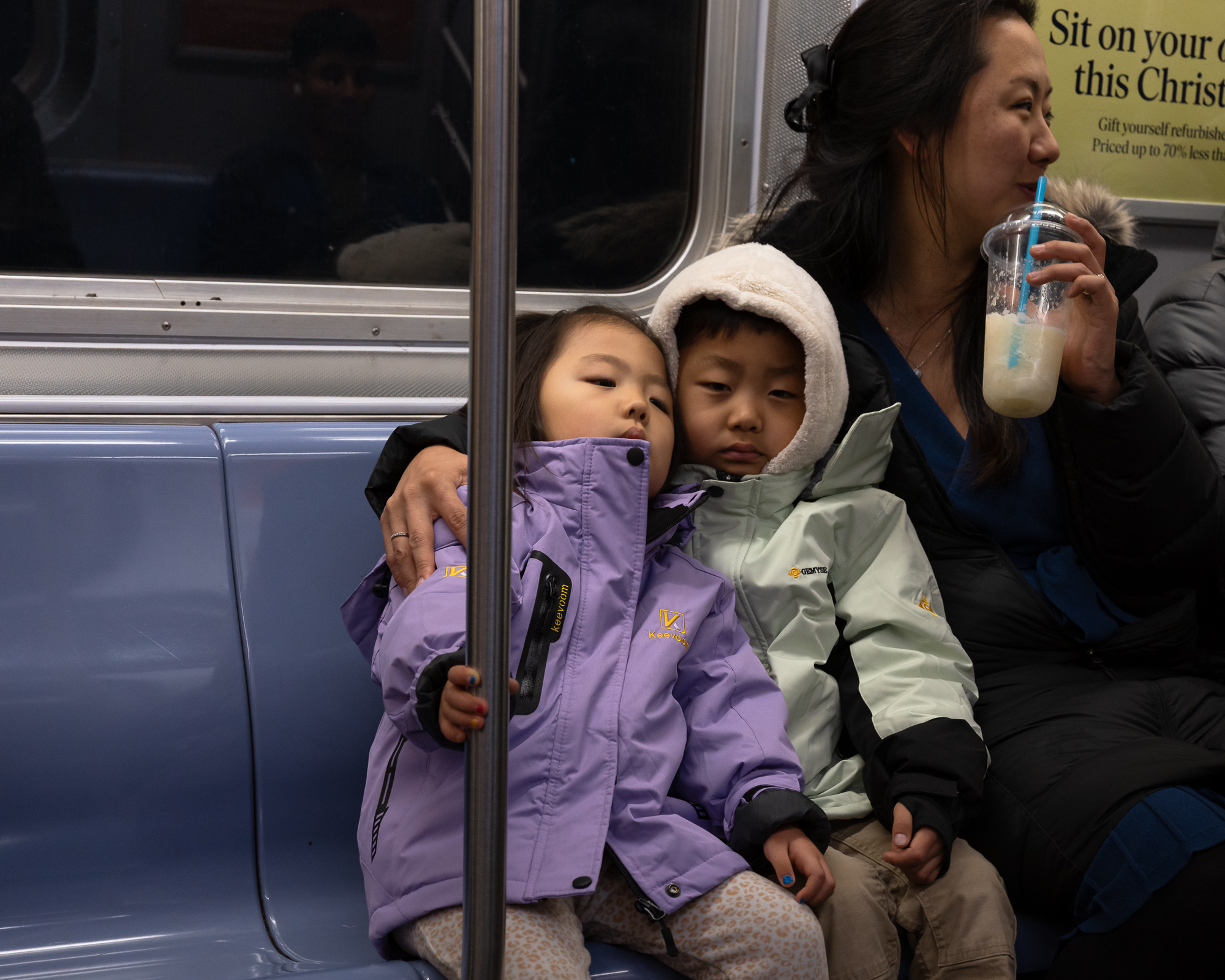 Two small children in puffy winter coats, one lavender and one pale mint with a fur-trimmed hood, sit pressed against each other on a blue subway bench while a woman beside them holds a plastic cup of iced drink with a blue straw to her mouth, one arm around the children.