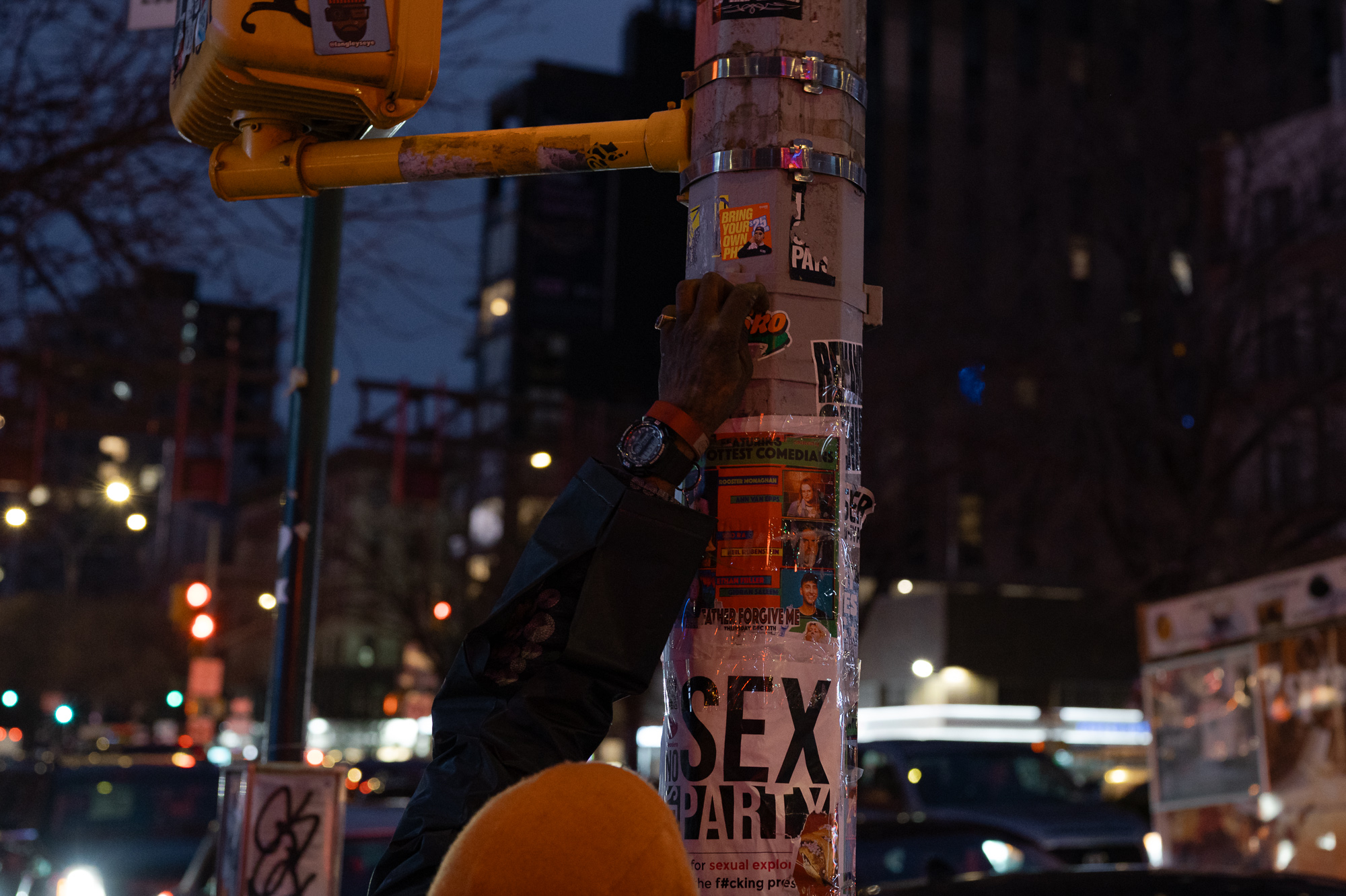 At dusk, a dark-sleeved arm with a black digital watch reaches up to pick at stickers on a metal traffic signal pole layered with handbills, including a taped poster reading SEX PARTY and a comedy show lineup. Behind it, a yellow pedestrian signal arm, blurred red and green traffic lights, and dim city buildings fill the blue evening sky.