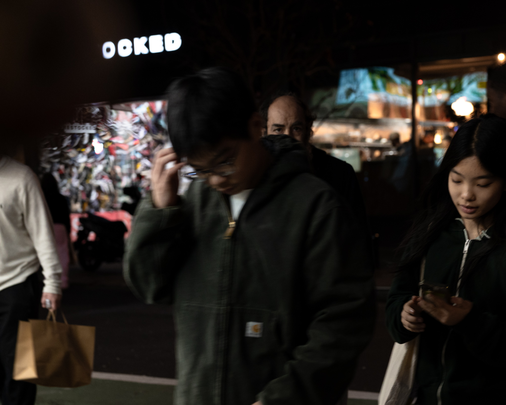 At night on a city sidewalk, a young person in an olive Carhartt hooded jacket pushes their glasses up while looking down, blurred in the foreground. Behind them a bald man in dark clothing meets the camera's gaze directly, and at right a woman in a dark zip-up hoodie looks at her phone, with a brightly lit sneaker store window and an illuminated sign reading part of the word 'DOCKED' in the background.
