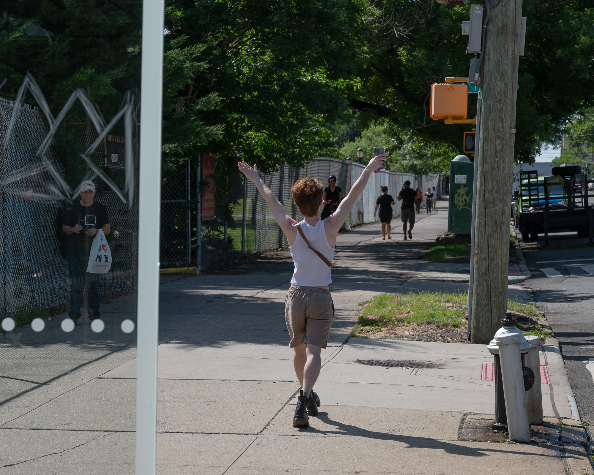 A person in a white tank top and tan shorts walks away from the camera on a sunlit sidewalk with both arms flung overhead, holding a phone aloft in one hand. Behind a glass panel at left, an older man in a baseball cap stands with an 'I Love NY' shopping bag, while pedestrians, a utility pole, a crosswalk signal and a fire hydrant fill the street beyond.