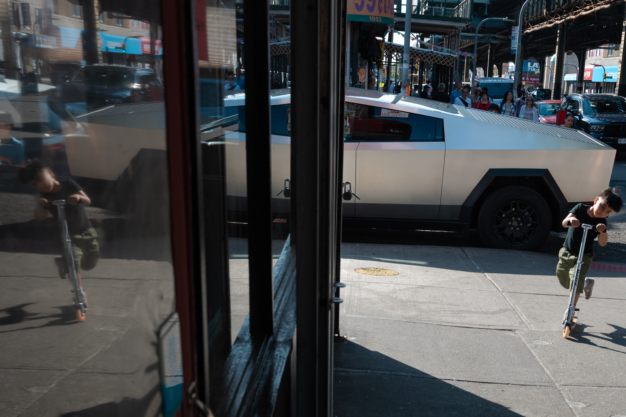 A boy in a black T-shirt and olive shorts pushes off on a kick scooter along a sunlit sidewalk, while his mirrored double appears in a storefront window at left. Behind him a silver angular Tesla Cybertruck is parked at the curb, with shoppers, awnings and the steel elevated train structure beyond.