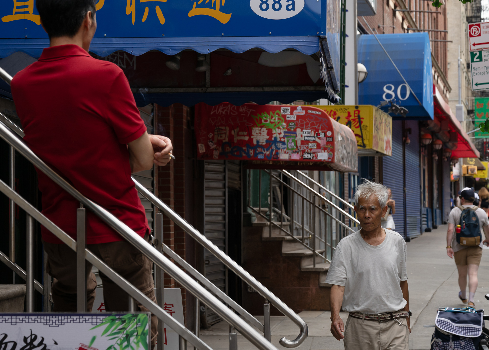 On a Manhattan Chinatown sidewalk, a man in a red polo shirt stands on a stairway behind a steel handrail with his back to the camera, holding a lit cigarette over the rail. Below and to the right, an older man with white hair in a pale T-shirt walks past shuttered storefronts under blue and graffiti-tagged awnings.