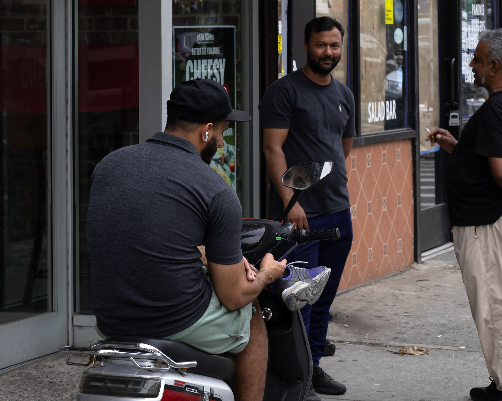 A man in a black cap, grey polo and mint green shorts sits sideways on a parked scooter outside a pizza shop, looking down at a phone mounted near the handlebars with a white earbud in his ear. Behind him a bearded man in a dark t-shirt and navy track pants stands on the sidewalk looking straight at the camera, while at the right edge an older grey-haired man in black holds a lit cigarette.