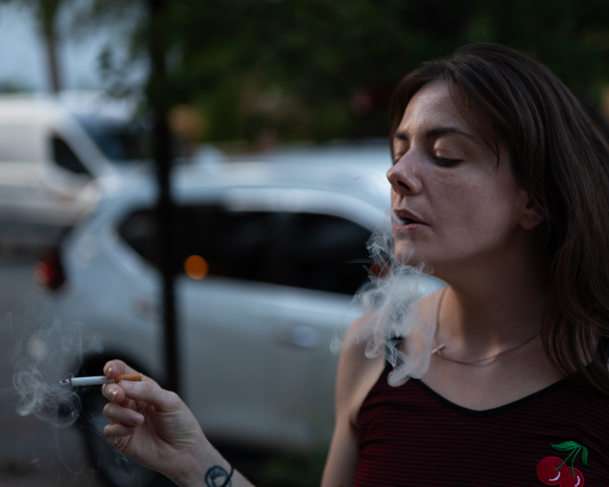 A woman with shoulder-length brown hair, eyes lowered, exhales a thick plume of smoke while holding a lit cigarette at chest height. She wears a red and black striped tank top with an embroidered cherry patch, and a blurred white car and tree fill the dim background.