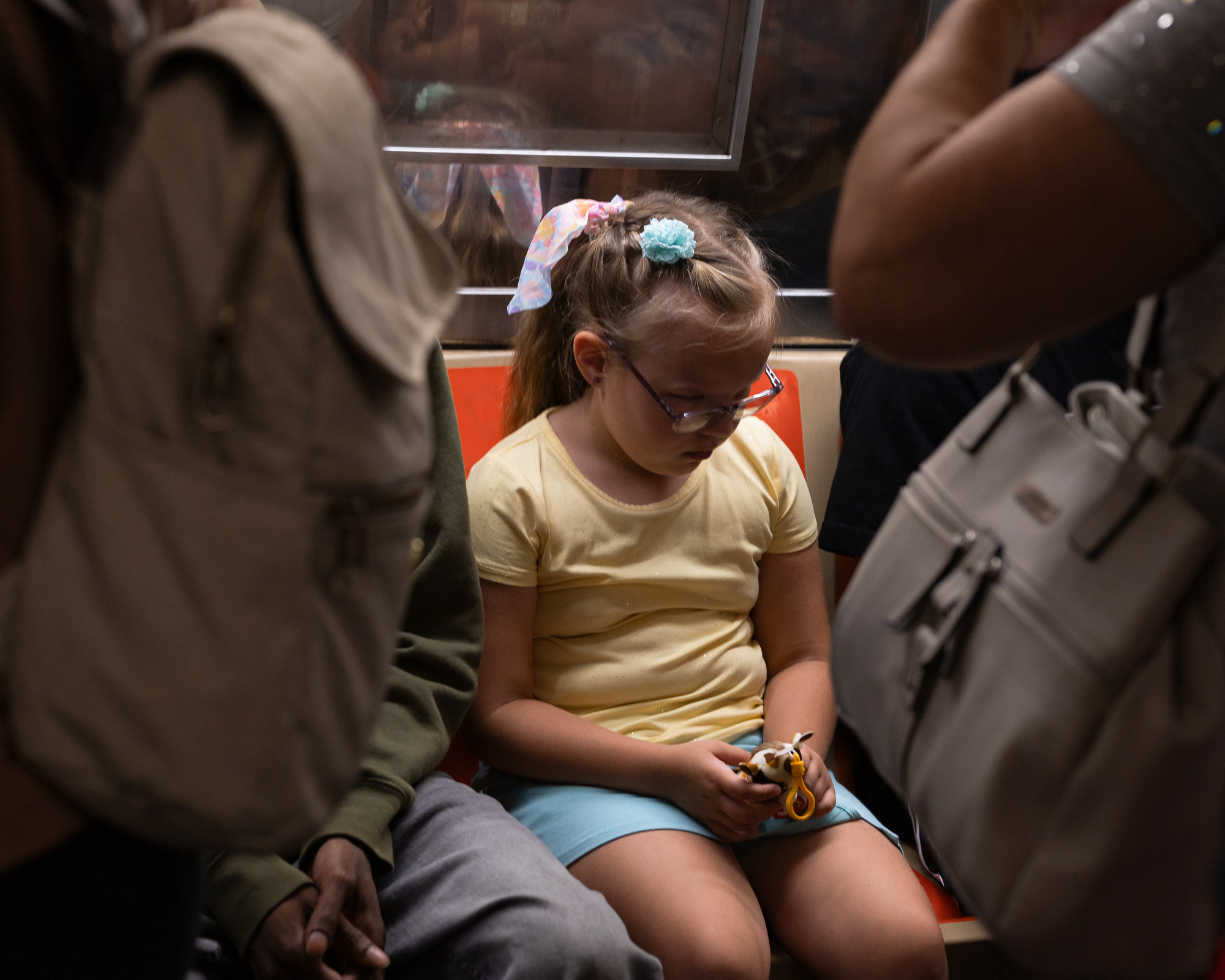 A young blonde girl in a pale yellow t-shirt and light blue shorts sits on an orange subway seat, head bowed, turning a small plastic horse toy in her hands. Standing passengers with backpacks frame her on both sides, blurred in the foreground.