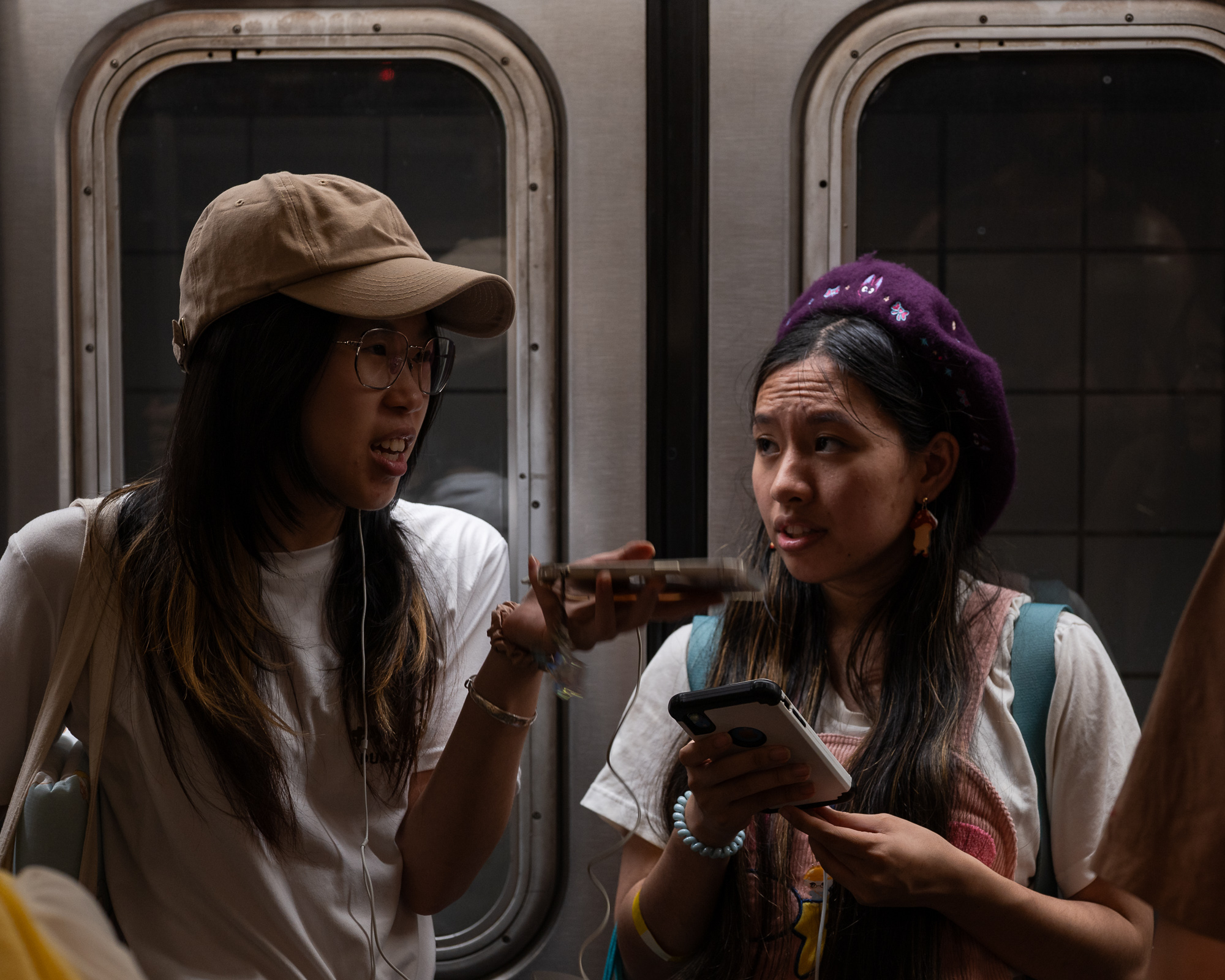 Two young women stand side by side in a subway car in front of closed doors, one in a tan baseball cap and wire-rimmed glasses talking with her hand raised, the other in an embroidered purple beret holding a phone in a white case and looking off to the side.