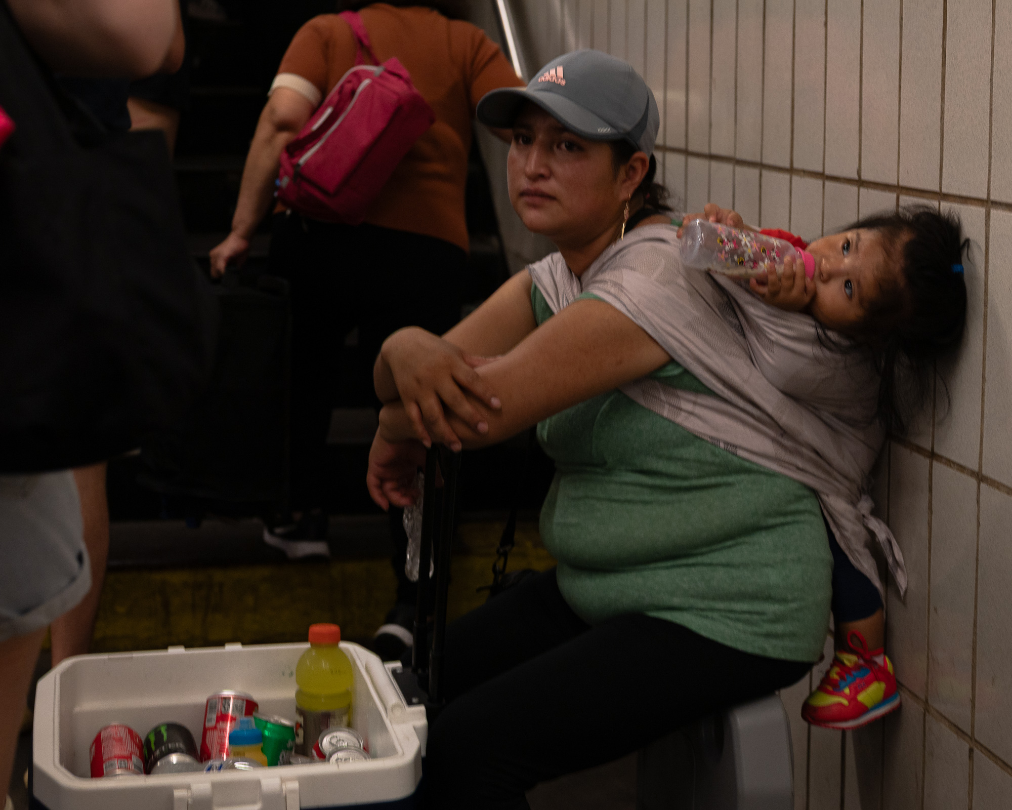 A woman in a grey Adidas cap and green shirt sits beside a white cooler filled with soda cans and a yellow sports drink in a tiled subway station, holding a bottle up to a young girl carried in a cloth sling on her back. Commuters climb a dim staircase behind them.