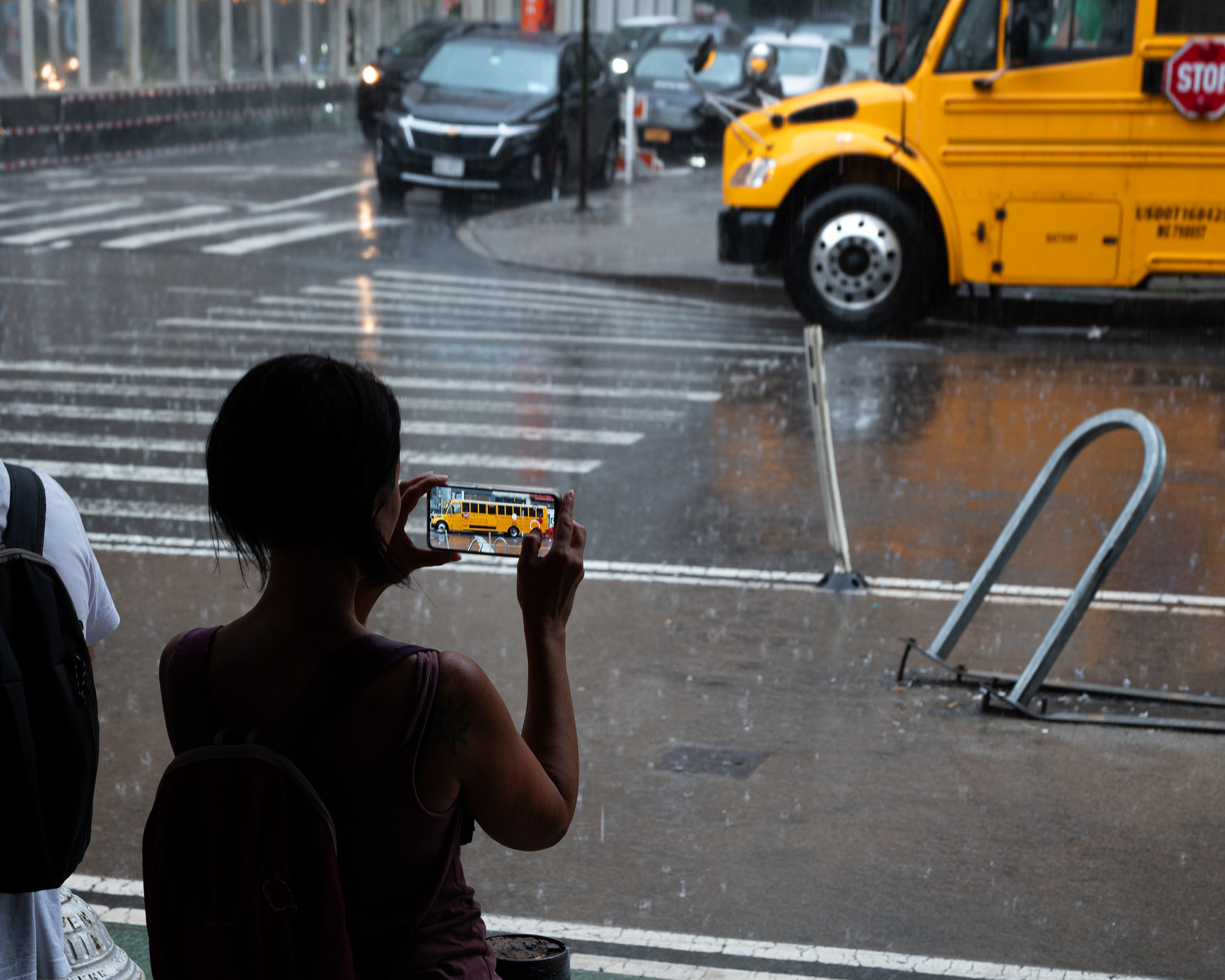 Seen from behind, a woman in a sleeveless maroon top holds up a phone, its screen showing a yellow school bus that sits across the flooded street in front of her. Rain streaks the intersection, a bent metal bike rack lies toppled on the wet sidewalk, and cars wait at the crosswalk in the gray light.