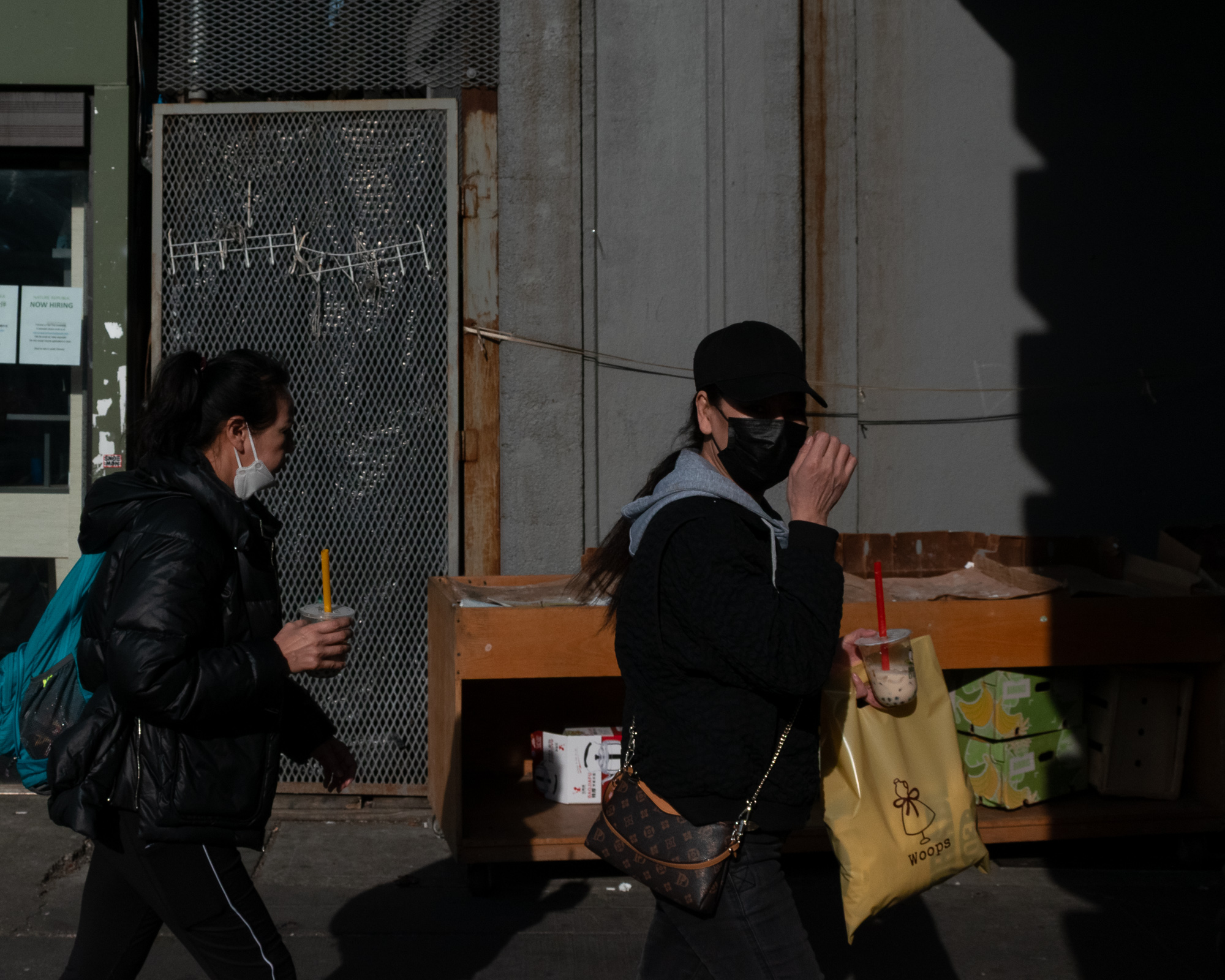 Two women in black jackets walk past a metal roll-gate and plywood hoarding on a New York sidewalk, each holding a clear cup of bubble tea with a wide straw. The woman on the right wears a black cap and black face mask and carries a yellow Woops shopping bag and a monogrammed shoulder bag; the woman on the left wears a white N95 mask and a teal backpack, her face lit by low sun.