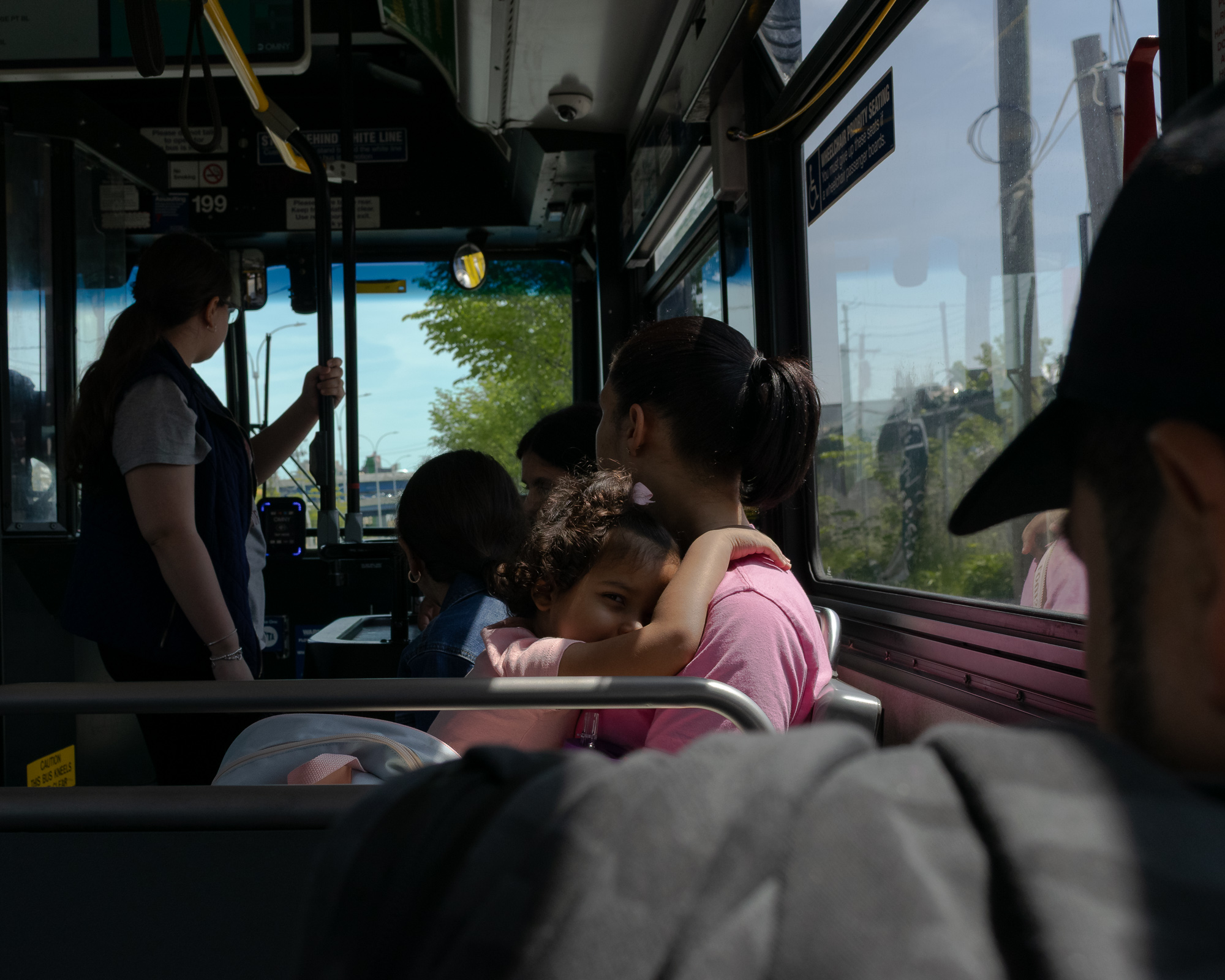 Inside a city bus, a young girl in pink rests her head on the shoulder of a woman with a dark ponytail, looking back toward the camera while sunlight falls through the window behind them. A standing passenger in a navy vest grips a pole near the front, and utility poles and green trees pass outside.