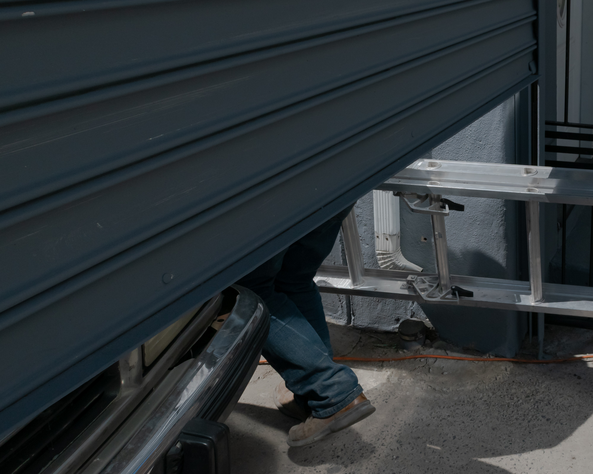 A partially raised blue-gray corrugated security shutter cuts diagonally across the frame, hiding everything above the waist of a man in jeans and worn sneakers who steps out from beneath it. To the right, an aluminum extension ladder leans against a painted concrete wall near a white downspout and an orange extension cord on the sidewalk.