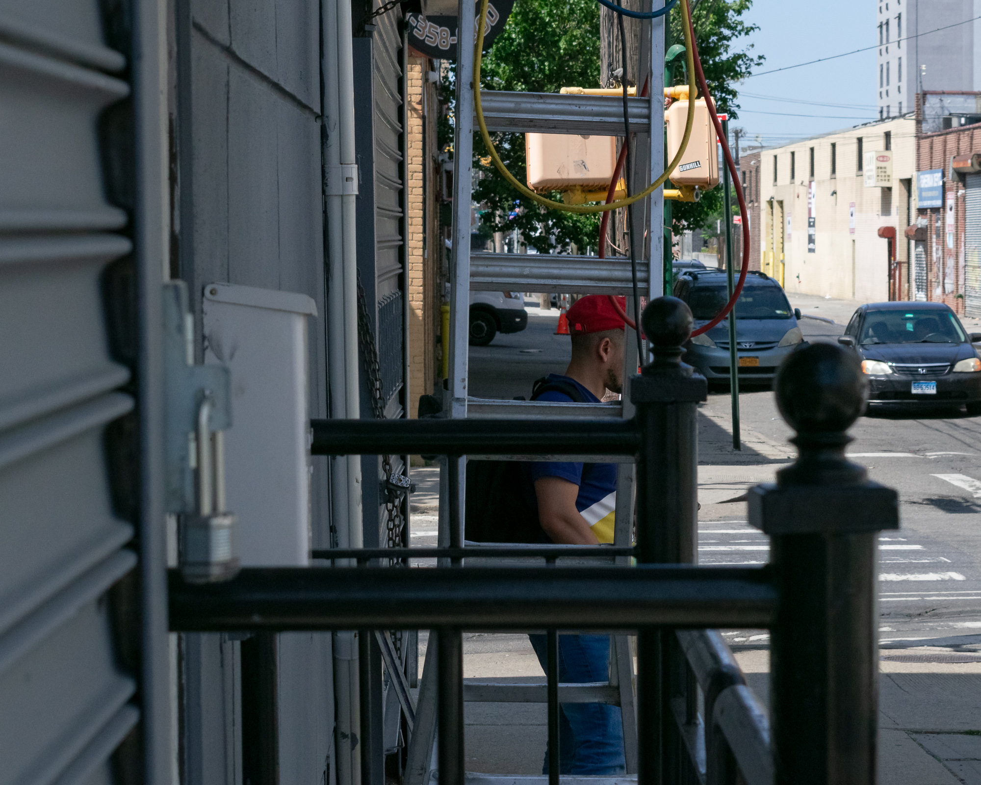 Viewed through a gap between a corrugated metal storefront and a black iron railing, a man in a red baseball cap and blue sleeveless shirt stands on a sunlit sidewalk, his body divided into sections by the rungs of a tall aluminum ladder. Behind him a parked minivan and a dark sedan sit along the curb beside beige industrial buildings.