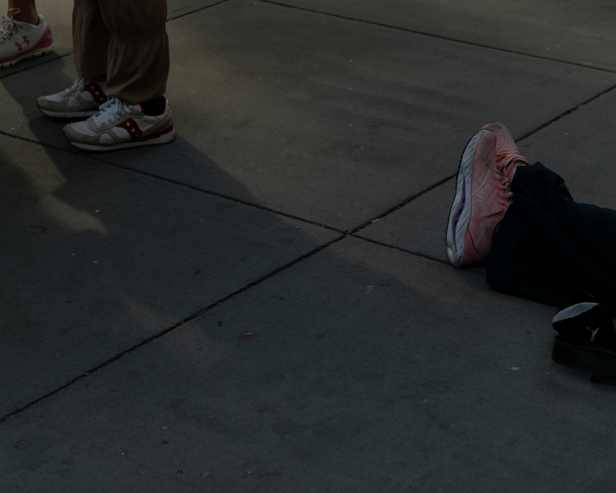 A low-angle view of a city sidewalk where sunlight cuts diagonally across the concrete slabs. At left, white and burgundy running shoes and a pink-trimmed sneaker stand near tan trousers; at right, a pink and purple running shoe and a dark loafer with a white sock rest at the edge of the light.
