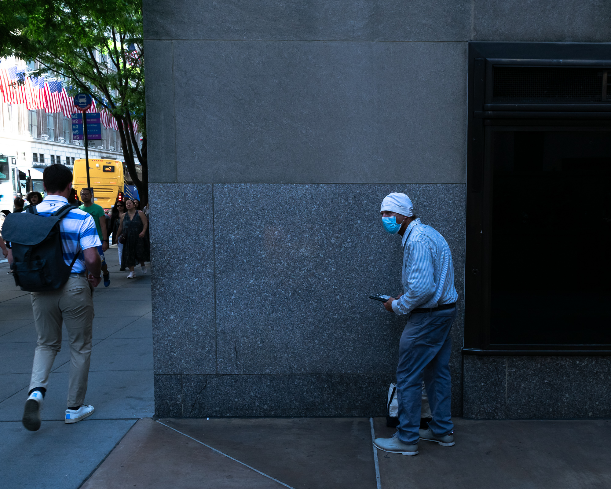 An older man in a white knit cap, blue surgical mask and striped shirt leans against a polished granite building corner in shadow, looking at a phone in his hand. Across the frame in daylight, a man in a striped polo with a grey backpack walks away past a yellow bus and American flags strung over the sidewalk.