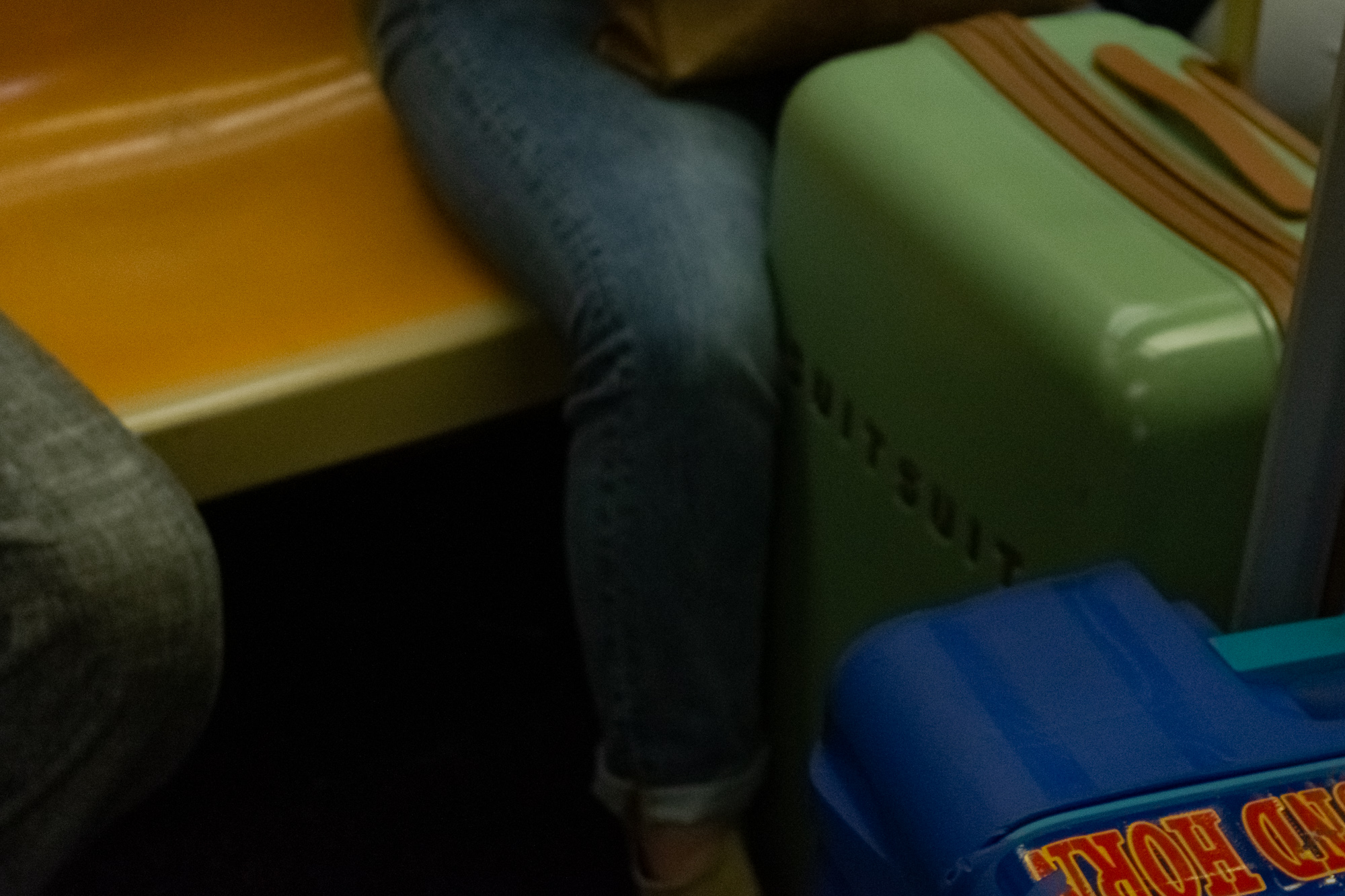 Interior of a New York subway car photographed from a seated passenger's eye level: a person in blue jeans sits with legs crossed on a mustard-orange molded bench, next to a mint green hardshell suitcase with a tan leather handle. A blue plastic bag with red lettering sits in the lower right corner, and the photographer's grey trouser leg fills the left edge.