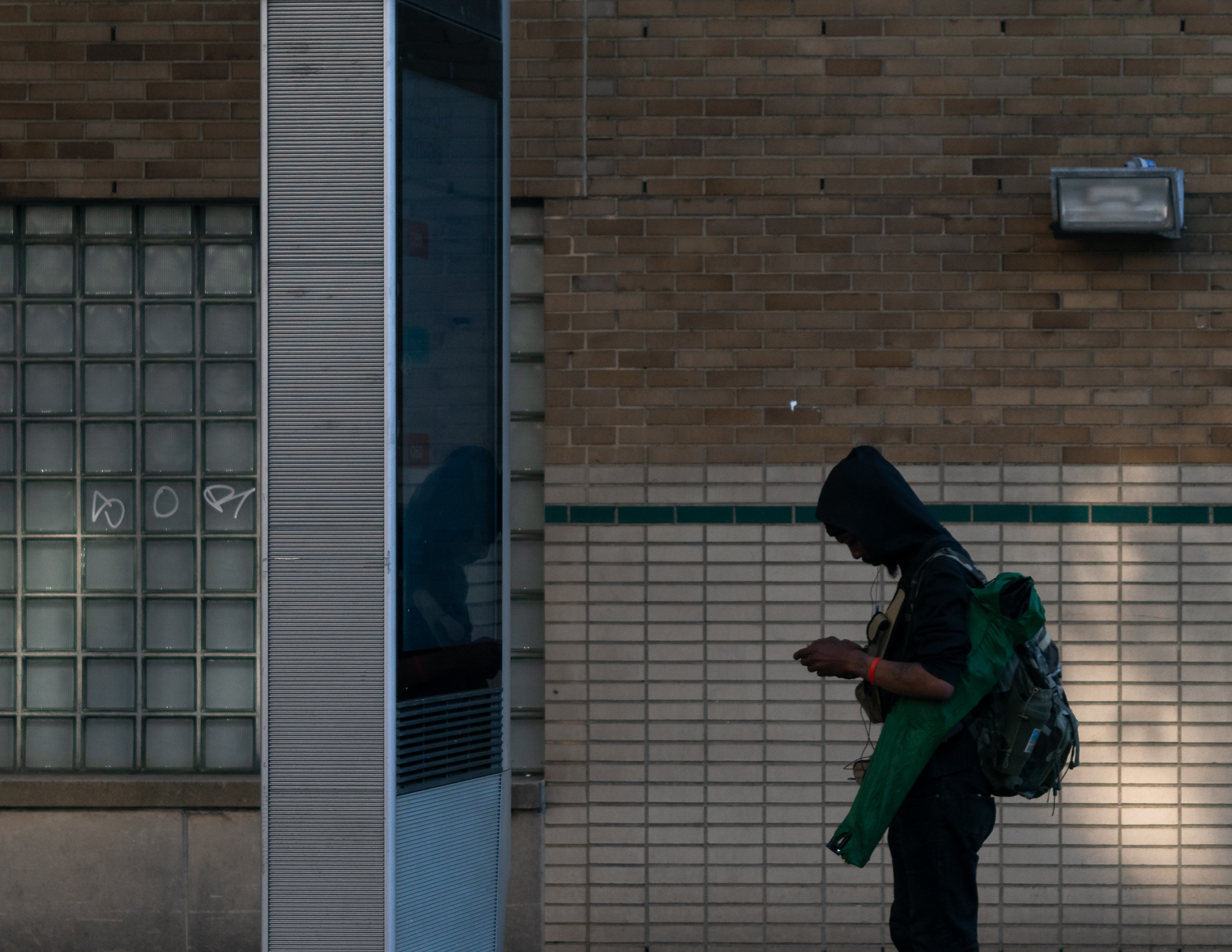 A person in a dark hooded sweatshirt stands in silhouette against a tan brick and tile wall, head lowered toward a phone held in both hands, with a green nylon jacket draped over one arm and a large backpack on their back. To the left, a glass block window with white graffiti and a narrow mirrored column reflect a dim figure.