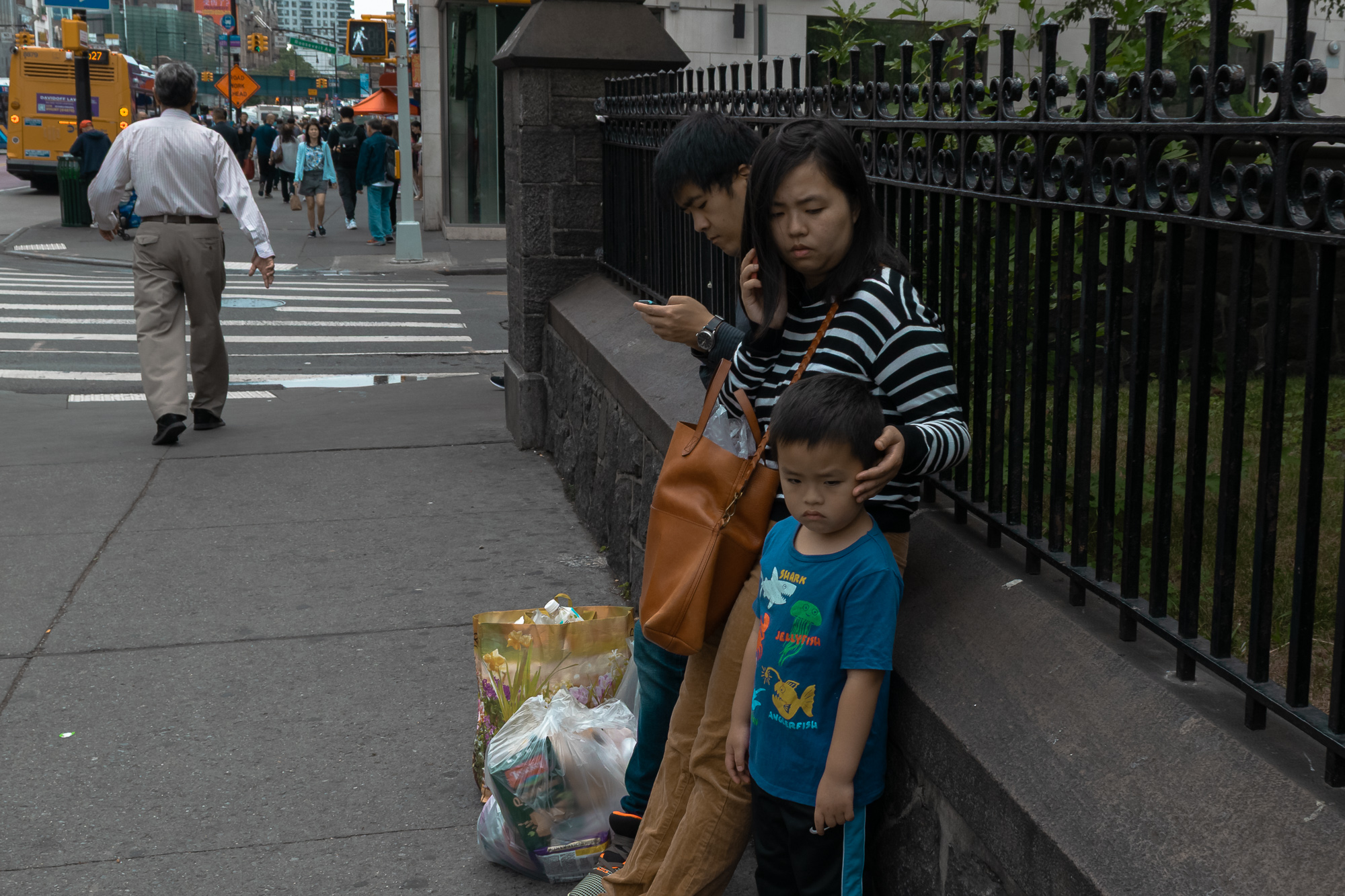 On a city sidewalk beside a black wrought iron fence and granite wall, a woman in a black and white striped sweater rests her hand on the head of a young boy in a blue T-shirt printed with a shark, jellyfish and anglerfish, while a man behind them looks down at his phone. Shopping bags of groceries and flowers sit on the pavement next to them, with a crosswalk and a yellow bus visible down the block.