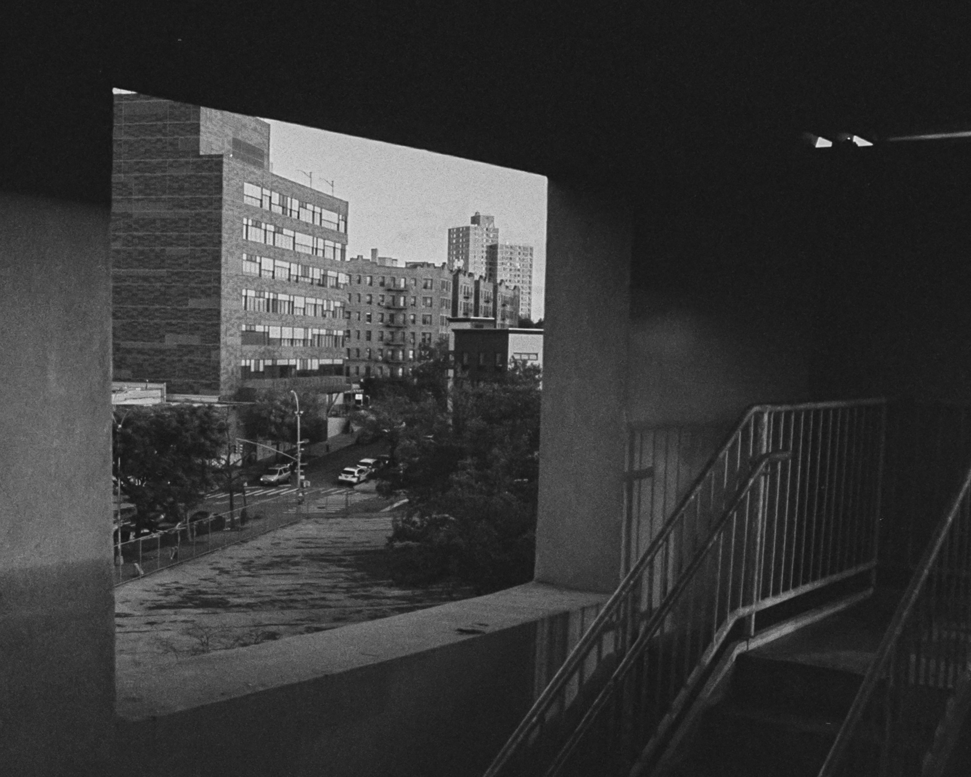 From inside a dark concrete parking structure, a large rectangular opening frames a bright view of a brick apartment block, mid-rise towers, street trees and parked cars below. A metal stair railing and a fluorescent light fixture sit in the shadowed interior at right.