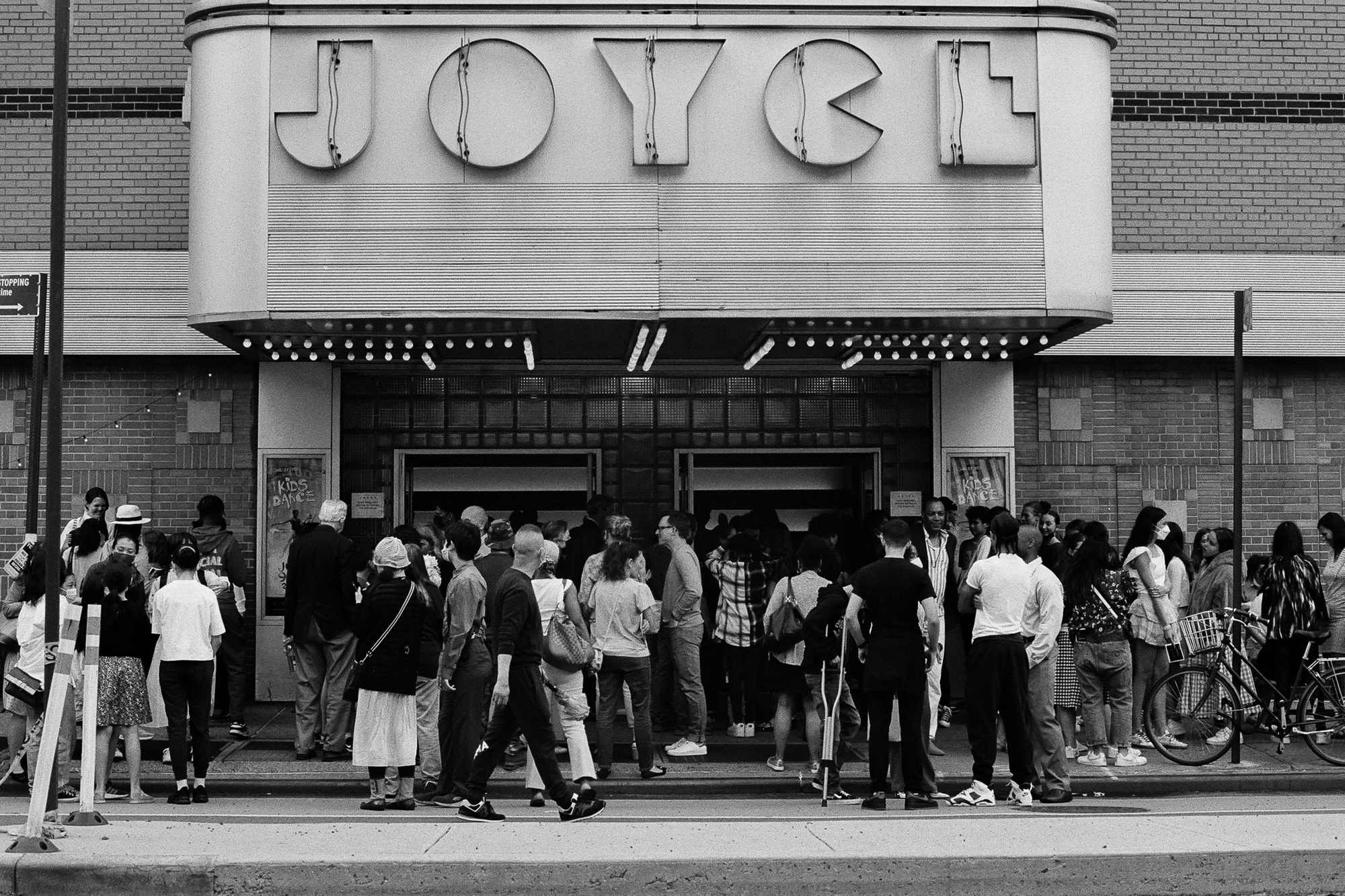 A black and white photograph of a dense crowd gathered on the sidewalk outside the Joyce Theater in Manhattan, standing beneath the curved Art Deco marquee with its large block letters and rows of bulbs. Two open doorways lead inside, flanked by posters reading Kids Dance, while a bicycle leans at the right edge of the frame.