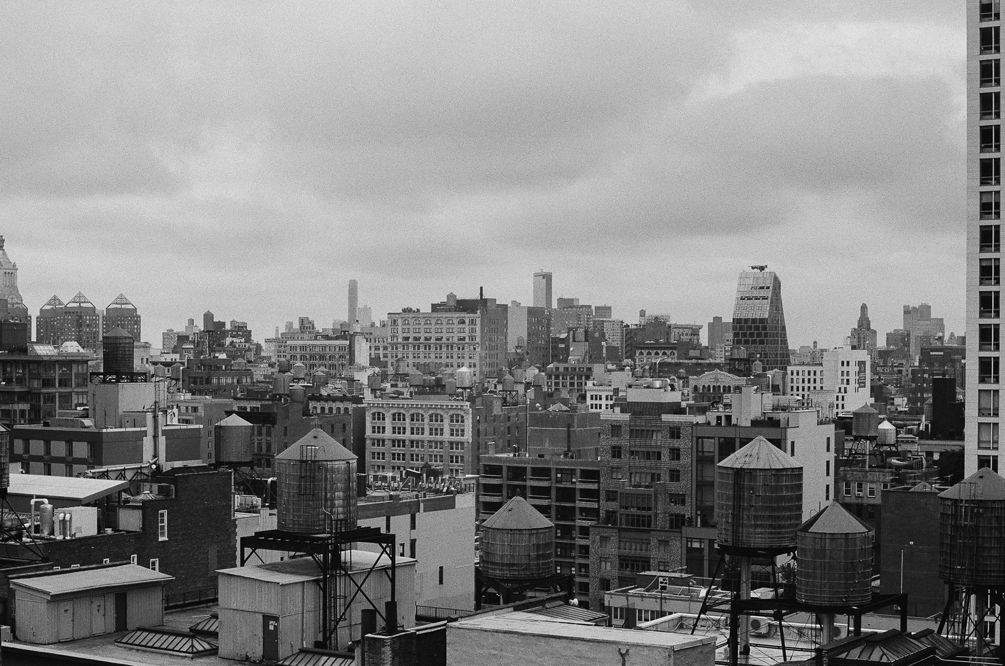 Black and white view across Manhattan rooftops, with a half dozen cylindrical wooden water towers on steel legs rising above tar roofs, skylights and brick parapets. A dense field of midrise buildings recedes into haze under a low, featureless overcast sky, with a tall apartment tower cropping the right edge of the frame.