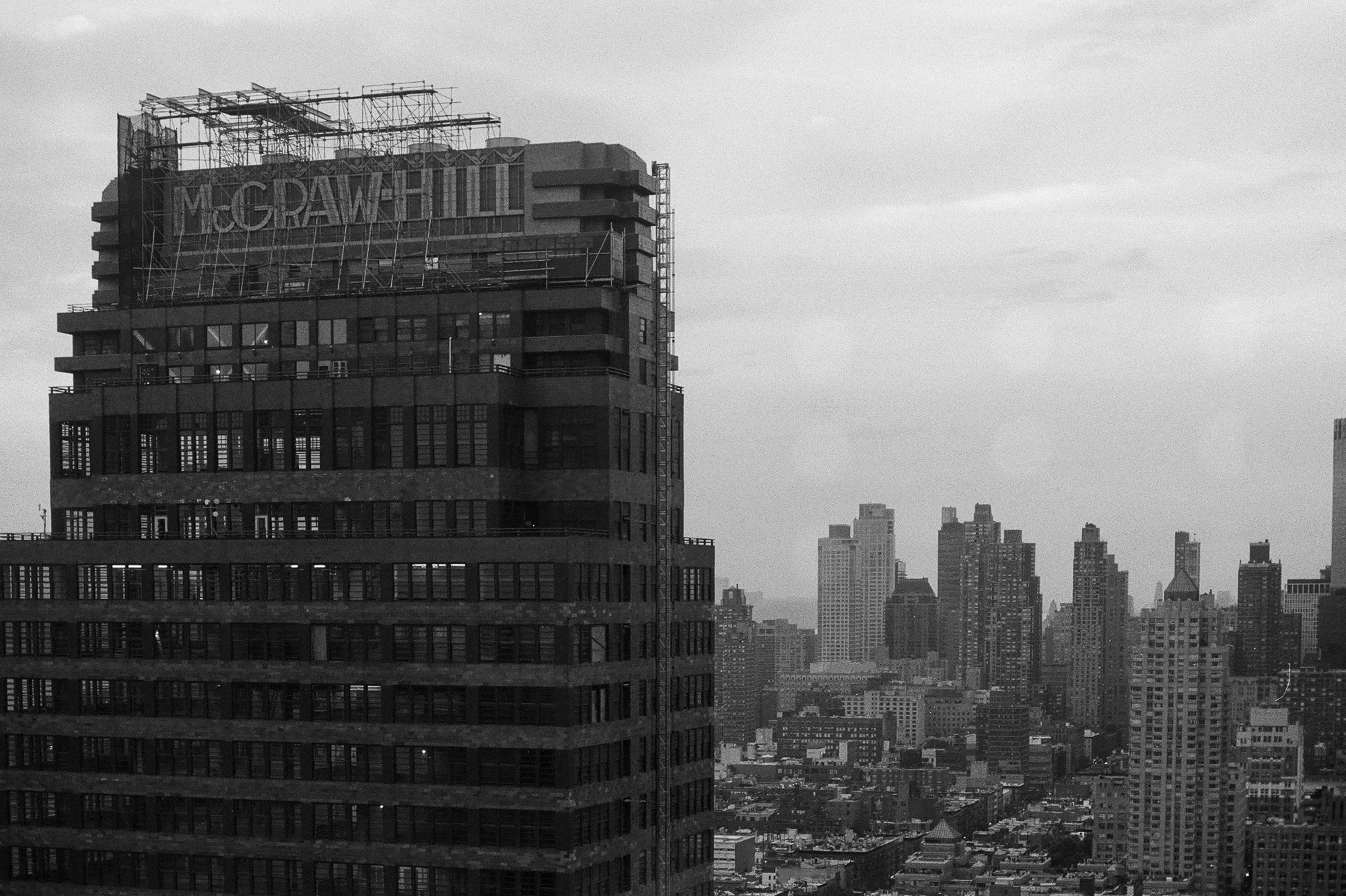 Black and white view from above of the McGraw-Hill Building in Midtown Manhattan, its rooftop sign wrapped in scaffolding and water tanks behind it, with hazy Hell's Kitchen towers receding into the distance at right.